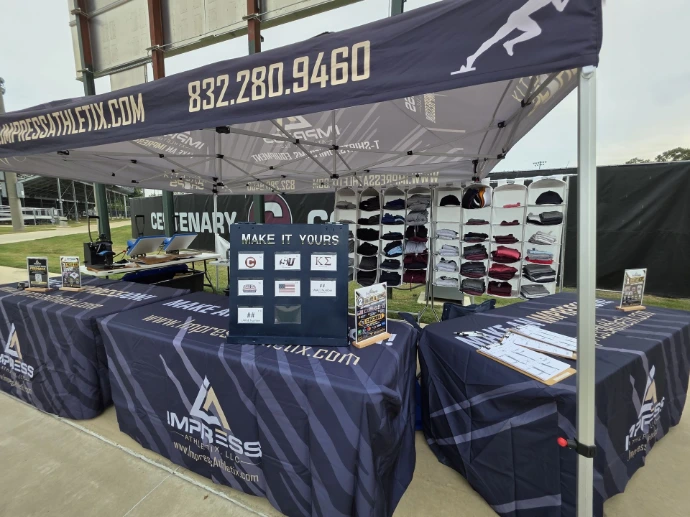 Impress Athletix event booth displaying apparel, hats, and a ‘Make It Yours’ customization display beneath a branded tent.