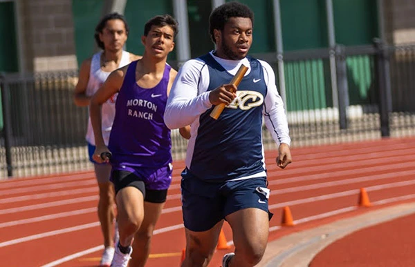 Three track athletes run on an outdoor track, with the athlete in front carrying a relay baton.