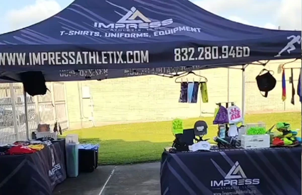 Impress Athletix vendor booth beneath a branded navy canopy at the Track Houston Championships, with apparel and track accessories displayed on tables.