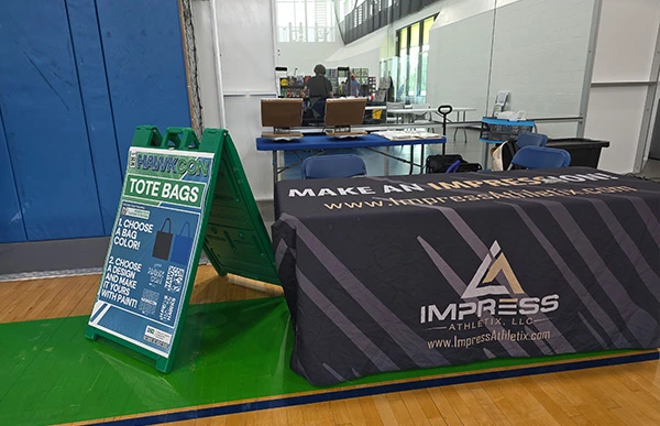 Impress Athletix’s HawkCon tote decorating station at UH-Clear Lake, with a sign advertising HawkCon tote bags.