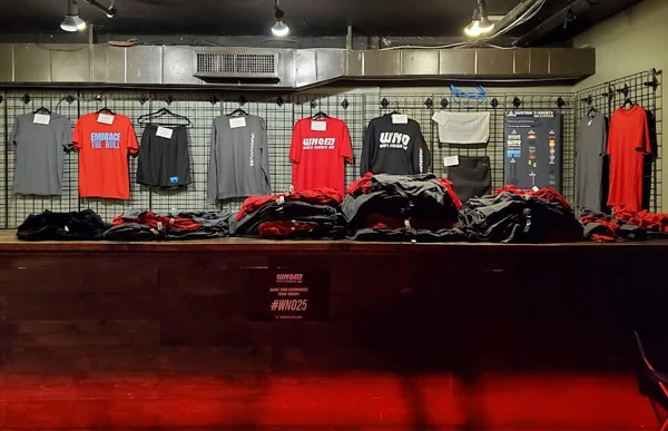 WNO 25 event apparel displayed on hangers and a counter at the Impress Athletix merchandise booth.