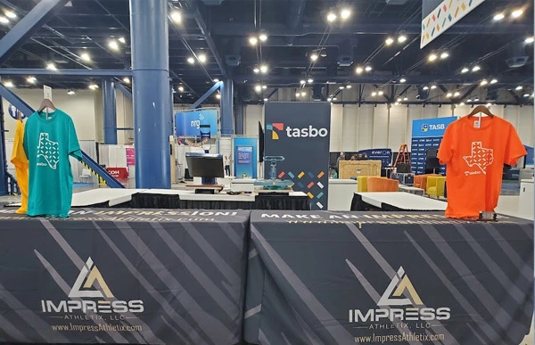 Impress Athletix booth at the 2024 TASBO event, displaying branded table covers and teal and orange shirts with a Texas design.
