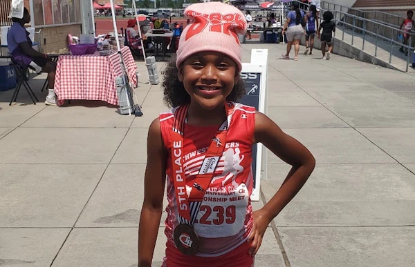 A young track athlete at the Track Houston Championships wears a pink “Throw Big” beanie and a medal.