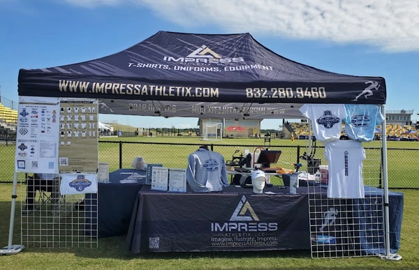 Impress Athletix booth at the 2023 SCAC Soccer Championships, with branded apparel and merchandise displayed beside a soccer field.