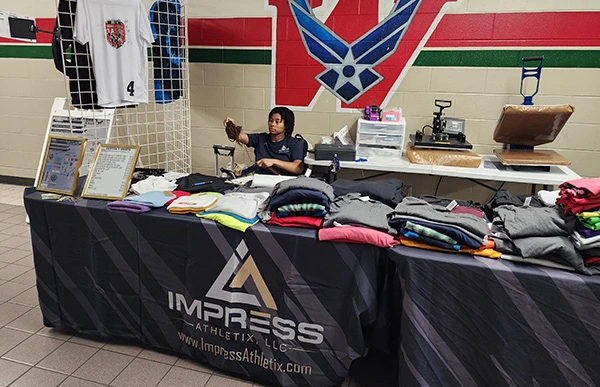 Impress Athletix booth at the 2024 Highlander Classic, with a tournament T-shirt displayed above a table of apparel.
